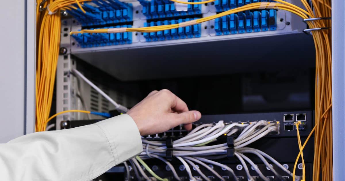 A person is reaching their hand toward a server rack with many organized yellow and white cables with blue connectors.