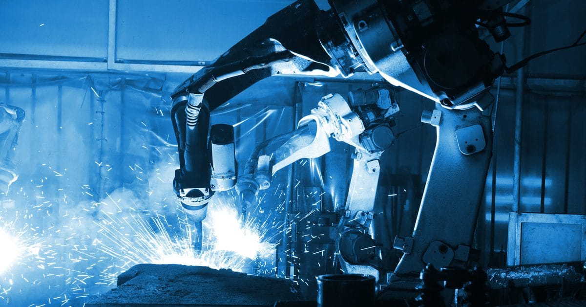A large automated welding machine in a dark factory welds a piece of metal as sparks fly up and around the workpiece.