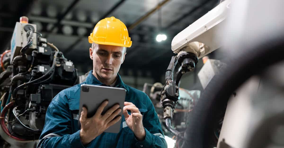 A man in a teal welding jacket and yellow hard hat holds a tablet. He’s in a factory with large machines behind him.