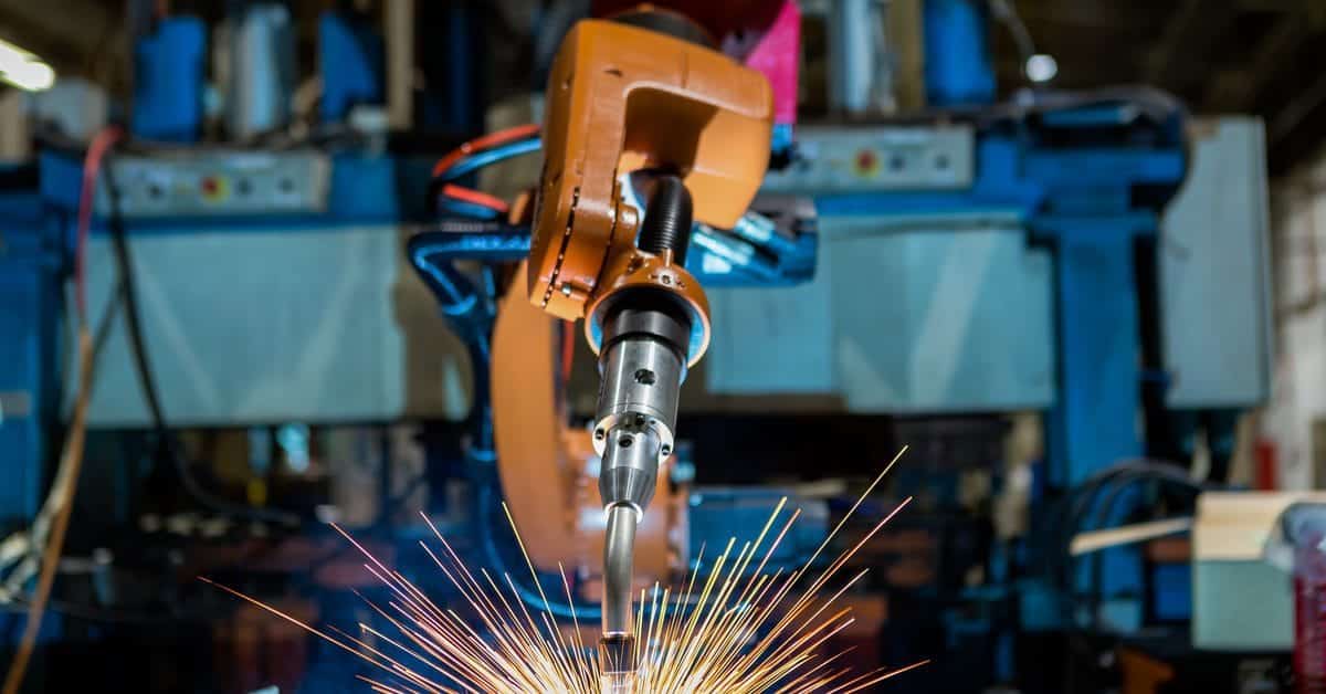 A large yellow robotic welding arm in a dark factory. Sparks fly up from the metal piece below the automated welder’s nozzle.