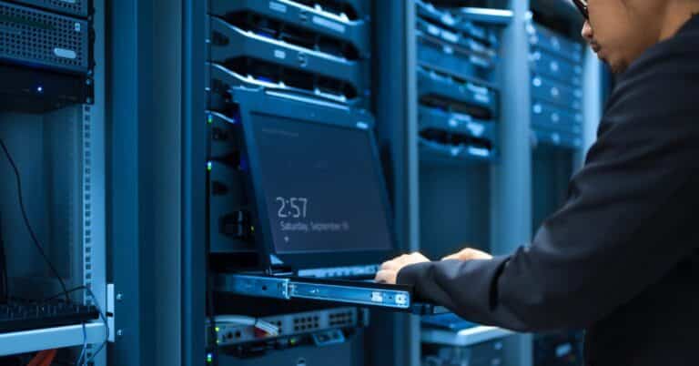 A man types on a laptop open to a screensaver resting on a pulled-out shelf in the aisle of a data center.