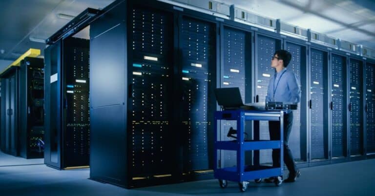 A man in professional attire walks past data server racks in a data center with a laptop on a blue cart.