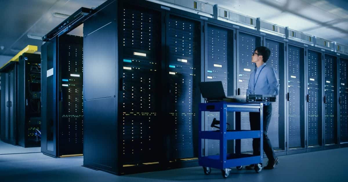 A man in professional attire walks past data server racks in a data center with a laptop on a blue cart.