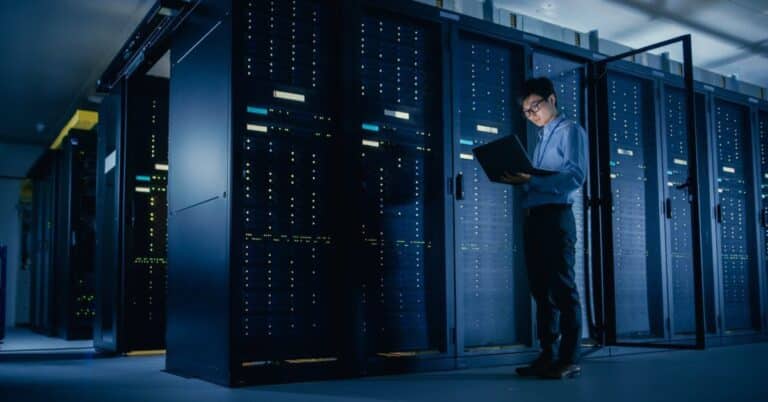 A man holds a laptop while standing in a dark room filled with data center racks that house servers.