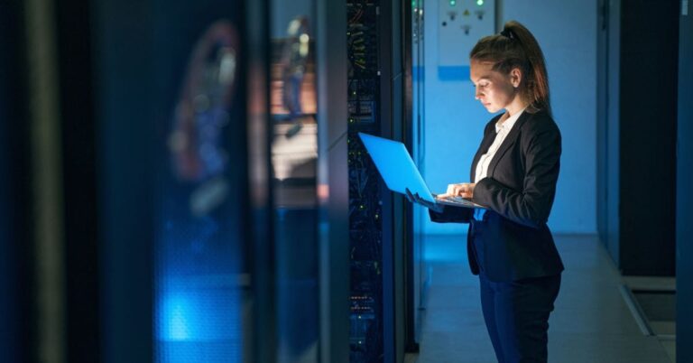 A woman with a high ponytail wearing a suit stands in a dark room in a data center and uses the laptop in her arms.