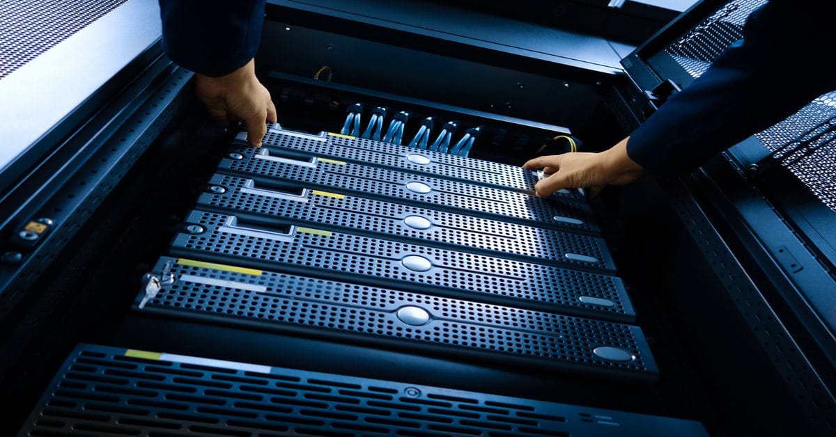 A pair of hands hold both sides of a server loaded onto a server rack under blue lighting in a data center.