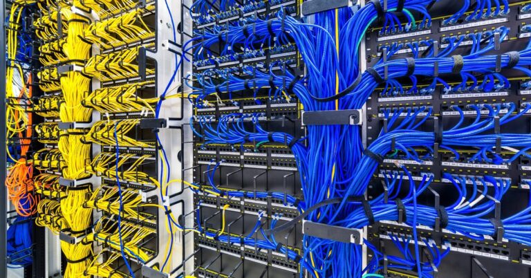 Bunches of blue and yellow cables are woven up a wall of a data center room, connected to server racks.