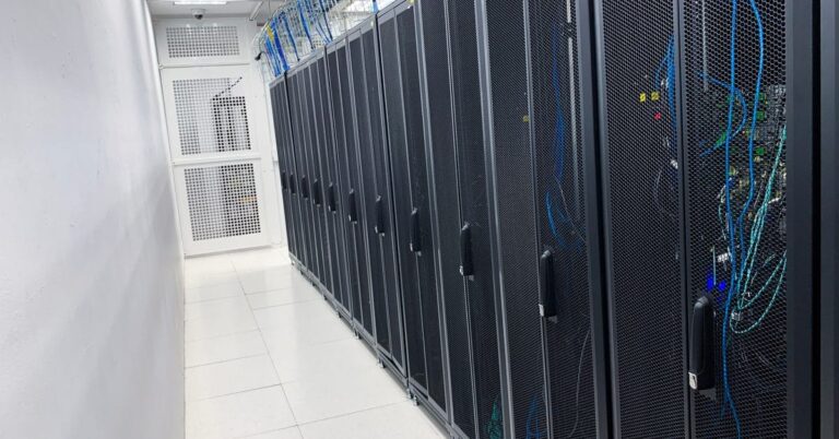 A row of black server racks stand inside a white room, stretching down on top of shiny tile flooring.