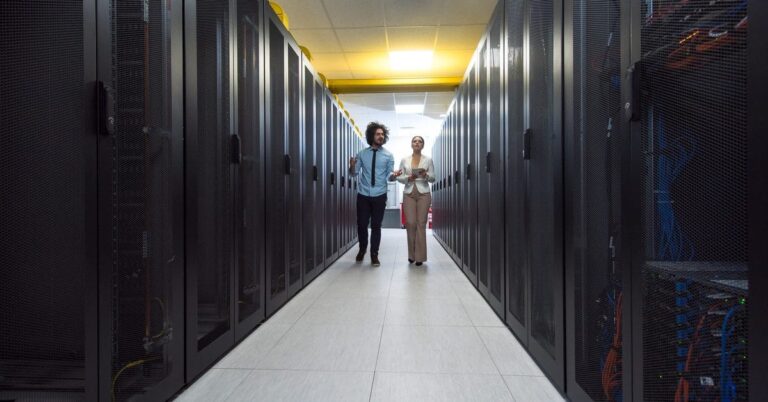 Two employees in professional attire walk down an aisle in a data center, with black server racks on both sides of them.