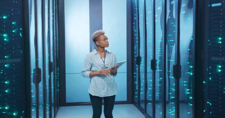 A woman stands in an aisle between rows of server racks on both sides. She holds a tablet against her side.