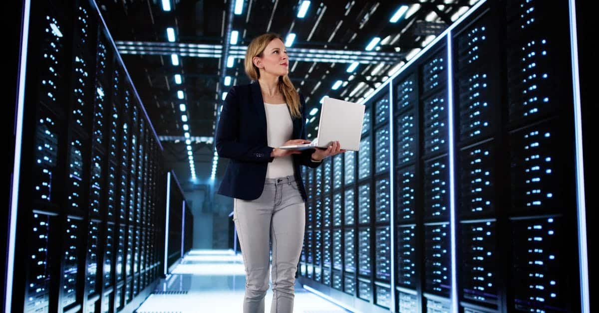 A woman in a professional jacket and holding a laptop walks through the aisle of a data center full of servers.