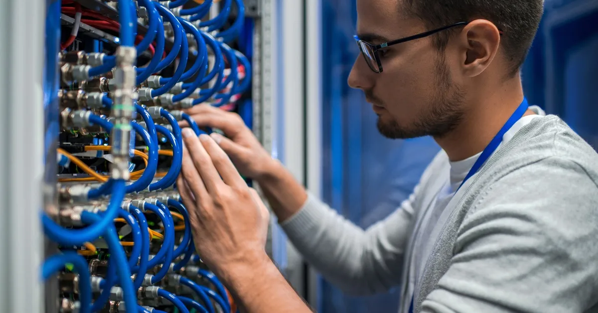 A man in a grey shirt and glasses stands in front of a data center network rack and messes with the blue cables.