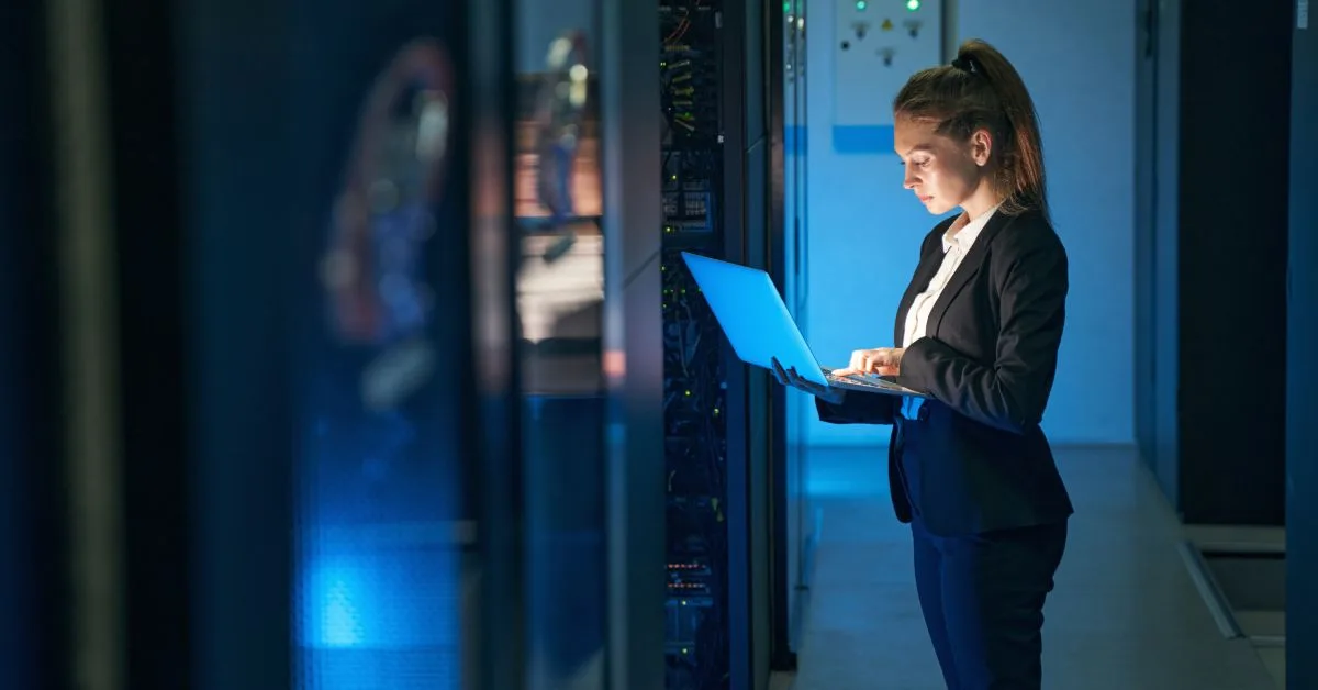 A woman with a high ponytail wearing a suit stands in a dark room in a data center and uses the laptop in her arms.