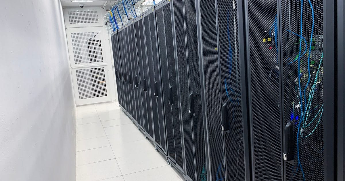 A row of black server racks stand inside a white room, stretching down on top of shiny tile flooring.