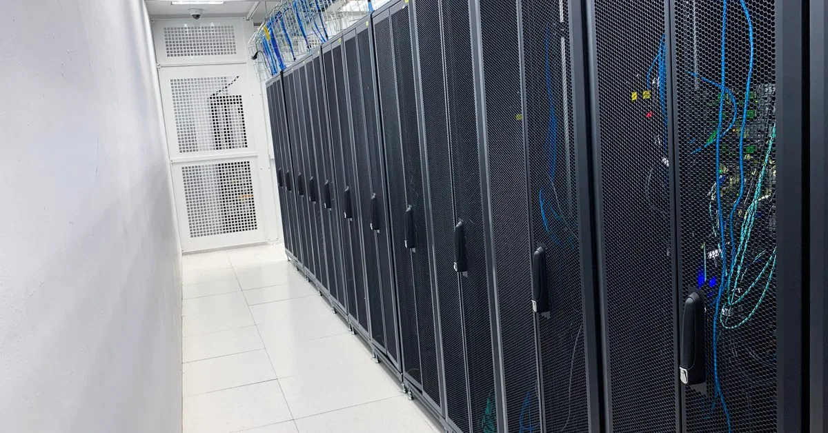 A row of black server racks stand inside a white room, stretching down on top of shiny tile flooring.