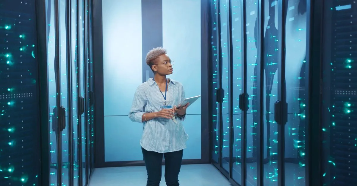 A woman stands in an aisle between rows of server racks on both sides. She holds a tablet against her side.