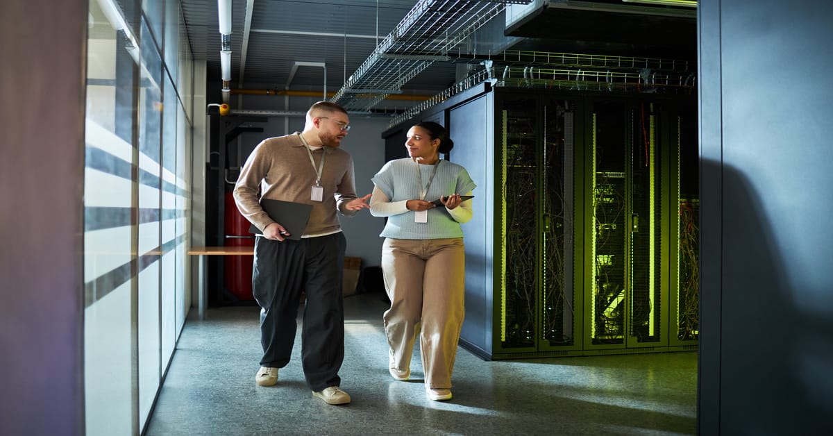 Two people wearing lanyards walk in a server room. There is a window on one side and server racks on the other.