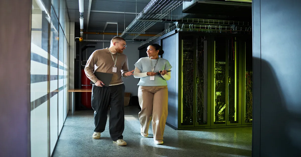 Two people wearing lanyards walk in a server room. There is a window on one side and server racks on the other.