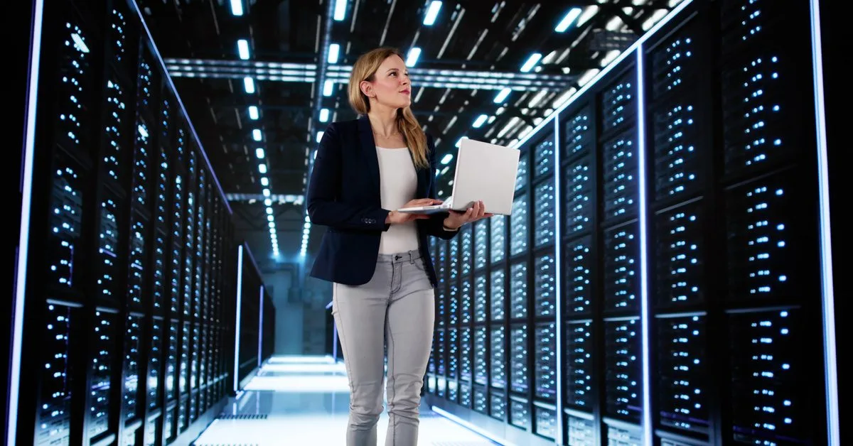 A woman in a professional jacket and holding a laptop walks through the aisle of a data center full of servers.