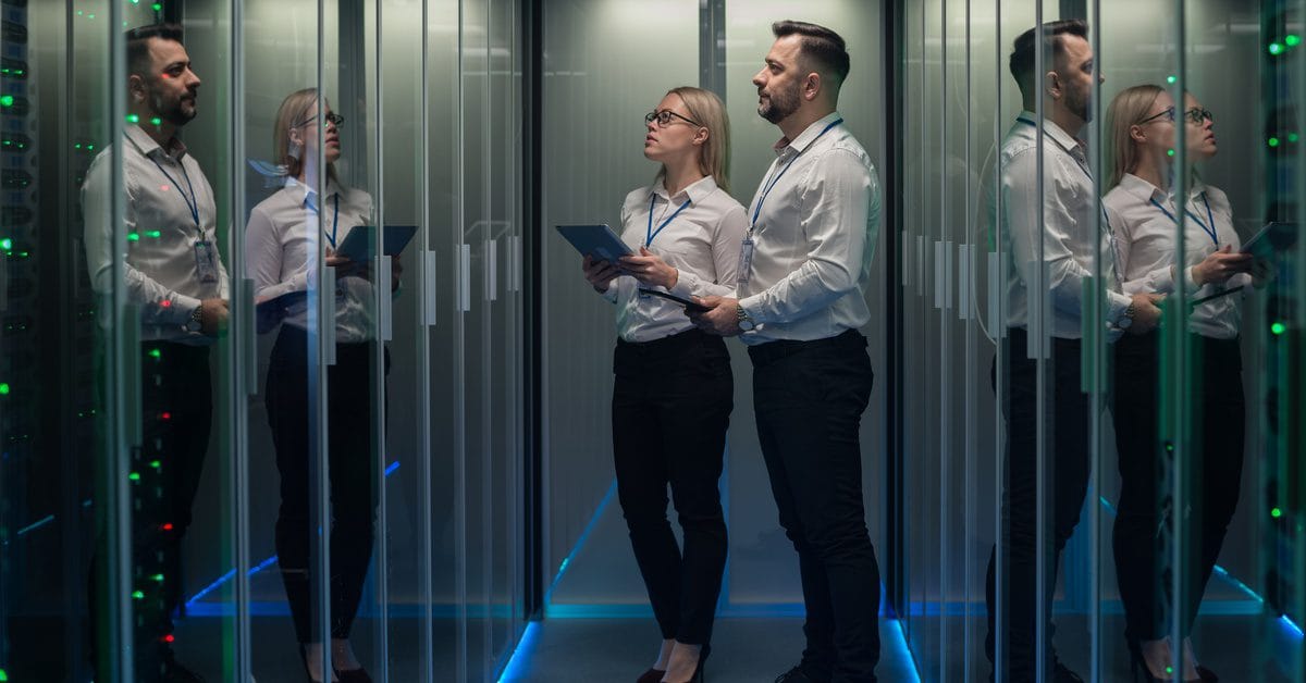 Two people wearing business attire stand in a narrow server room. Both are holding tablets and looking up at a server rack.