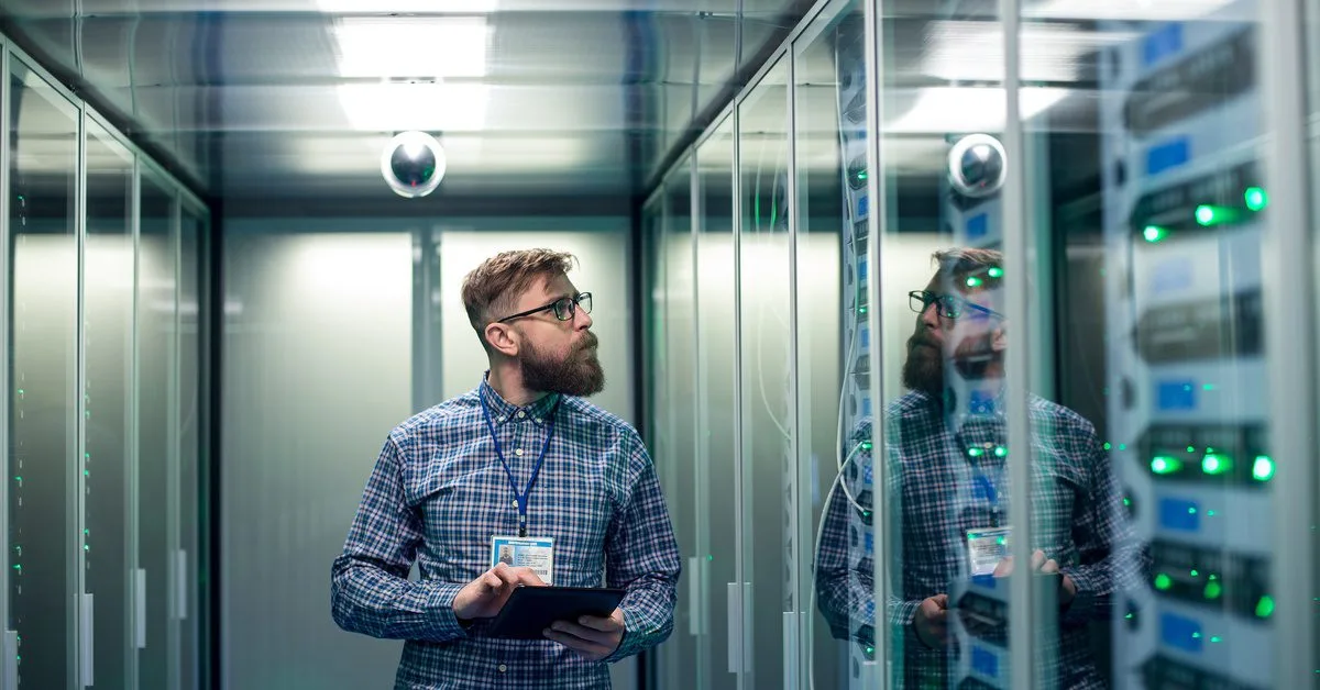 A man with a beard holds a tablet and looks intently at a server rack behind glass doors in a server room.