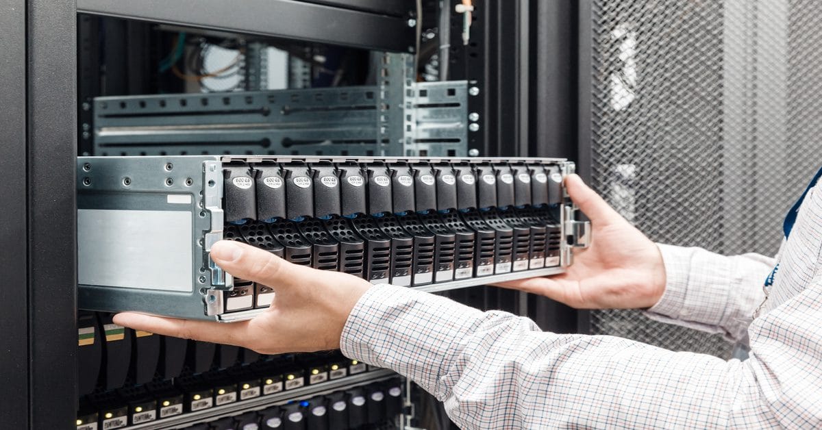 A person inserts a silver storage enclosure with black drive caddies into a server rack in a bright data center.