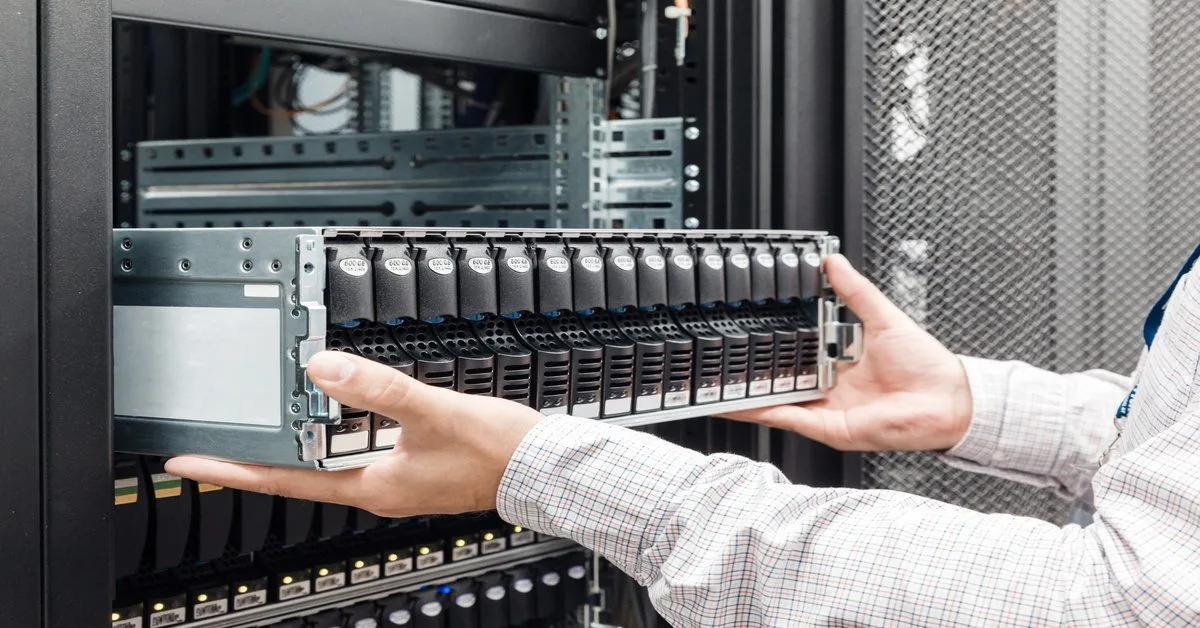 A person inserts a silver storage enclosure with black drive caddies into a server rack in a bright data center.