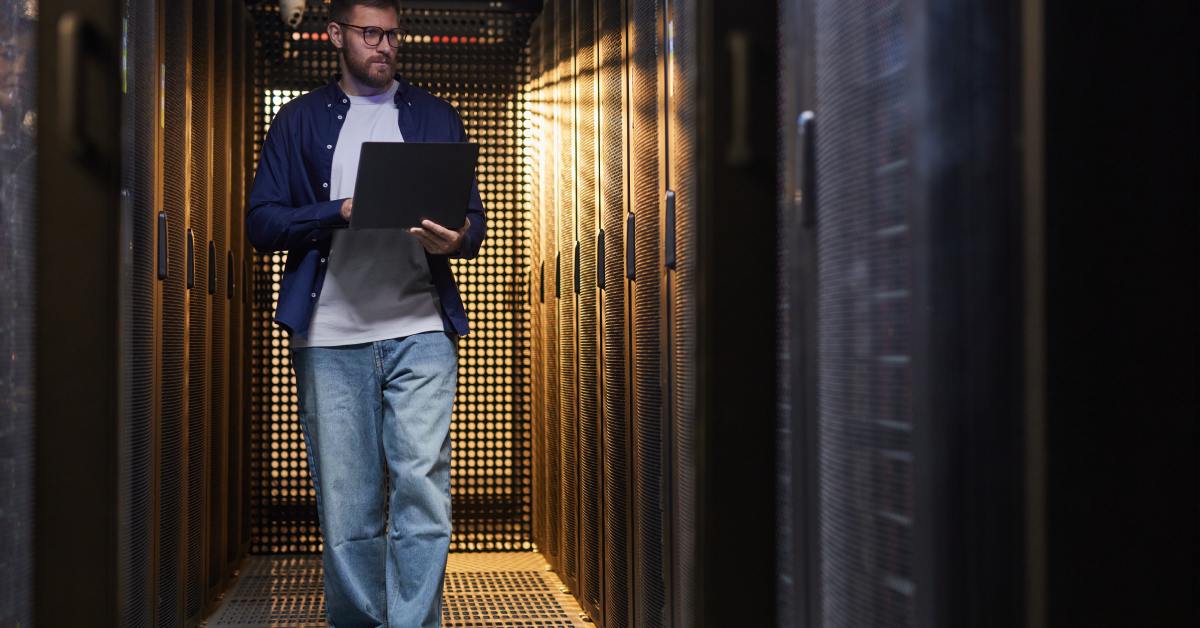 A person wearing glasses walks down the center of a data center aisle. He is holding an open laptop.