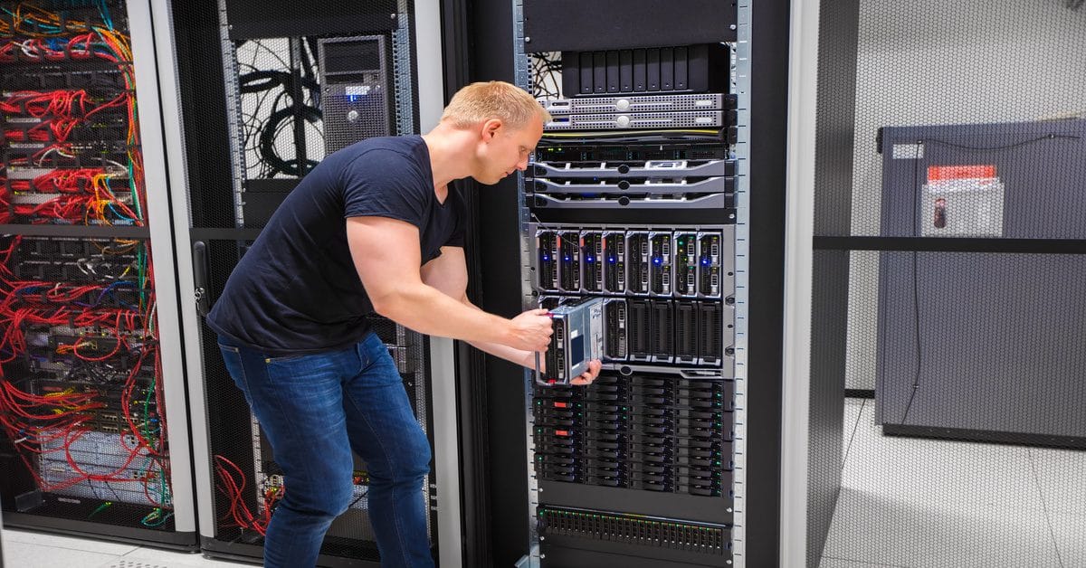 A technician installs a server into a metal rack inside a data center with visible cabling and network equipment.